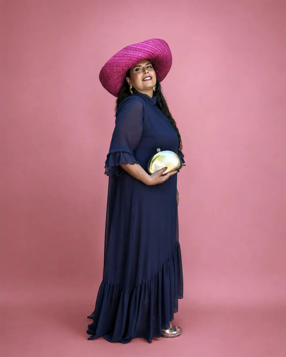 Vestido Marina de gasa azul marino con bajo de volantes, vista de perfil con pamela fucsia de ala ancha y clutch dorado, talla grande